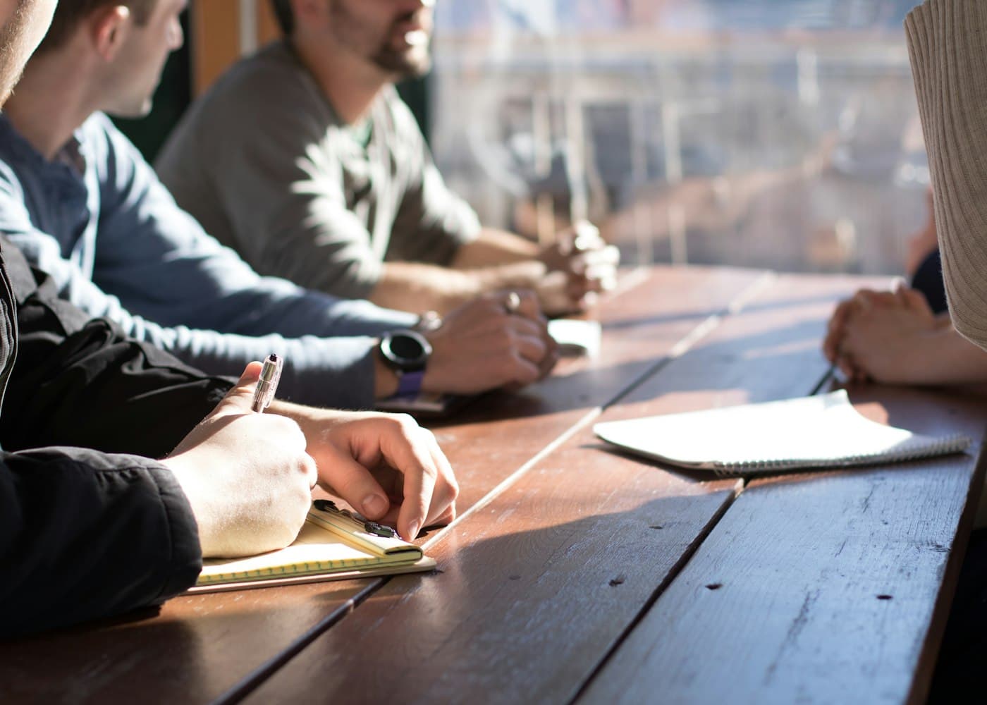 People sitting along a sunlit wooden table in conversation.